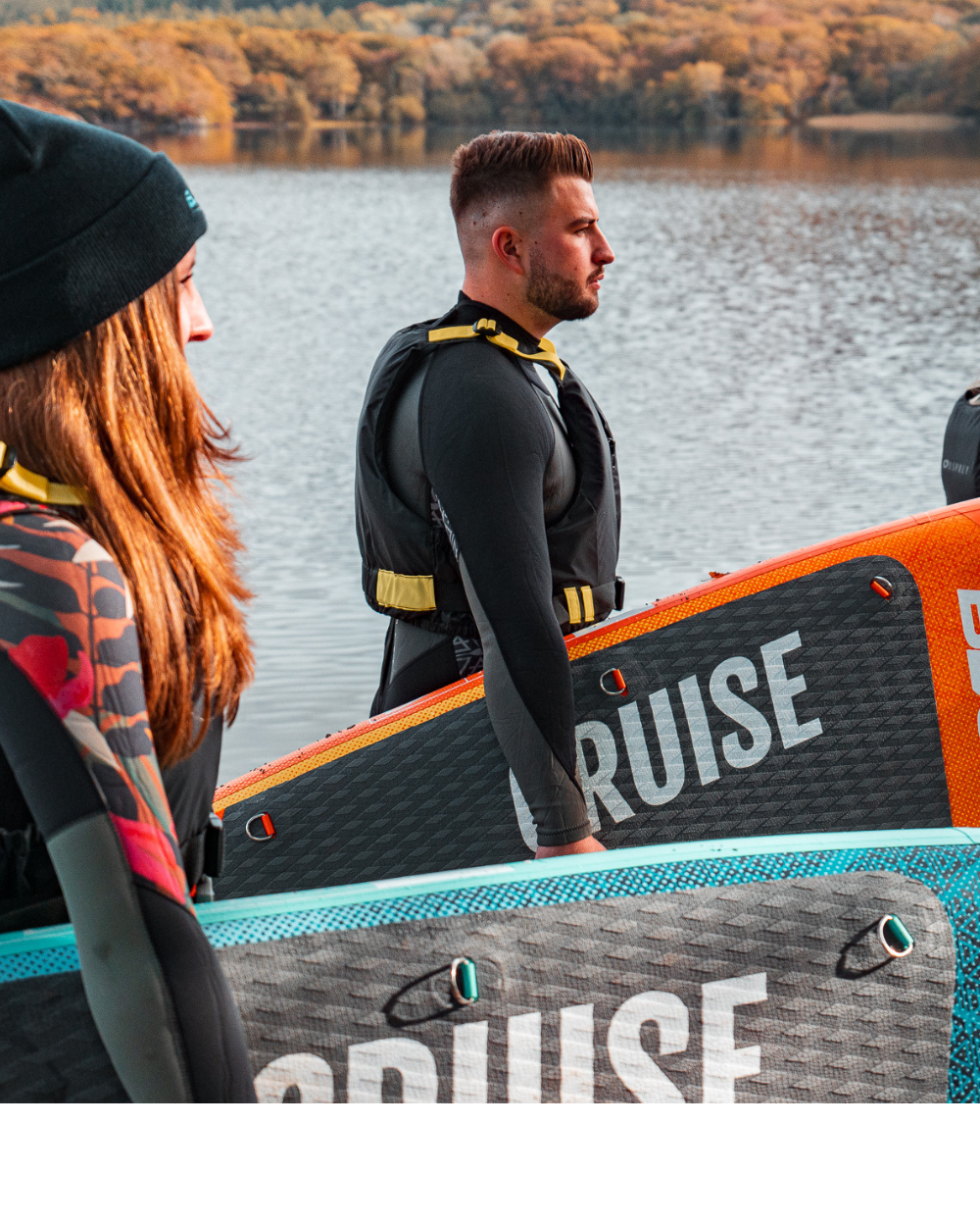 Two people in wetsuits holding inflatable paddleboards by a lake, autumn foliage in background