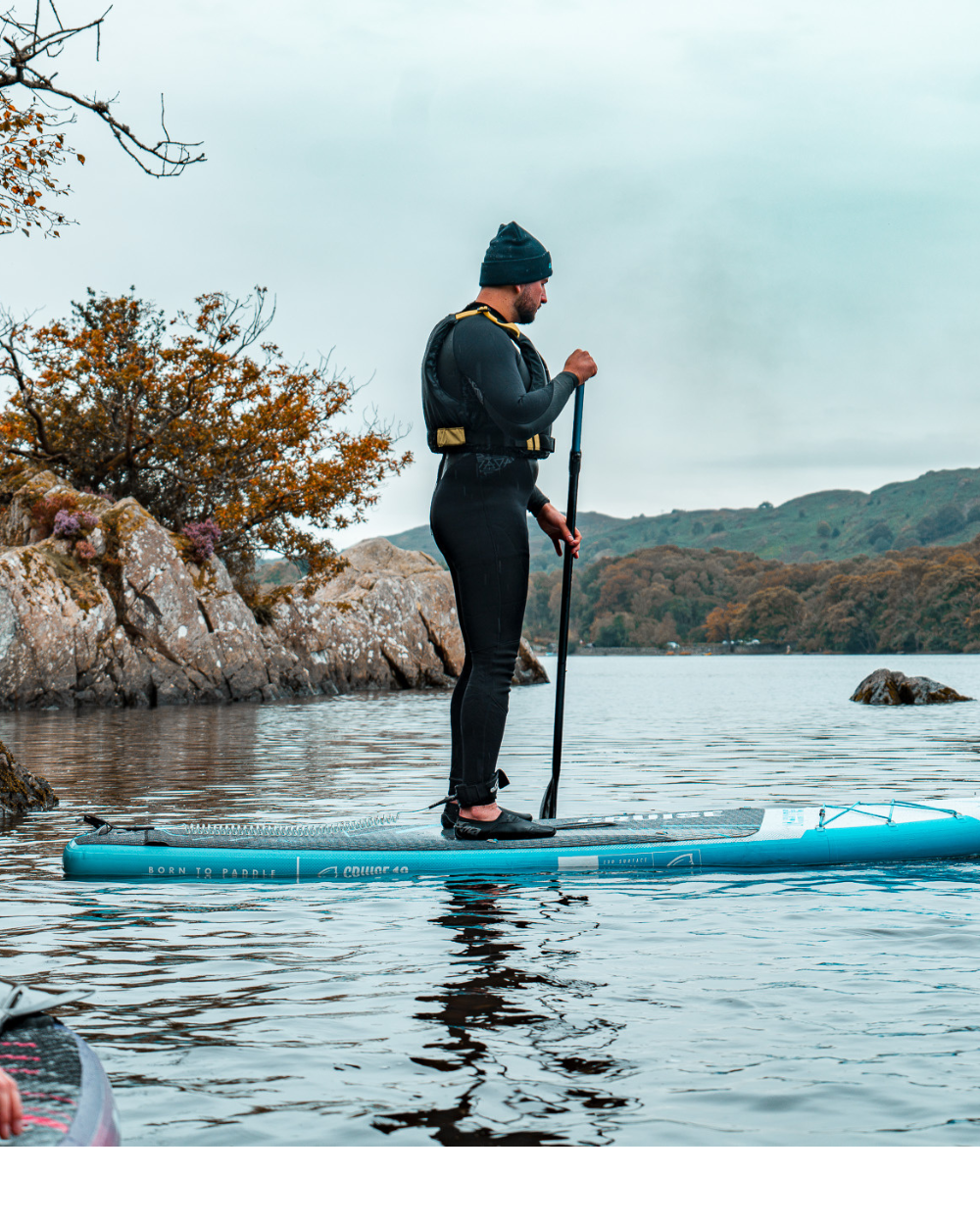 Person in wetsuit paddling on a blue inflatable paddleboard in calm water with rocky shore.
