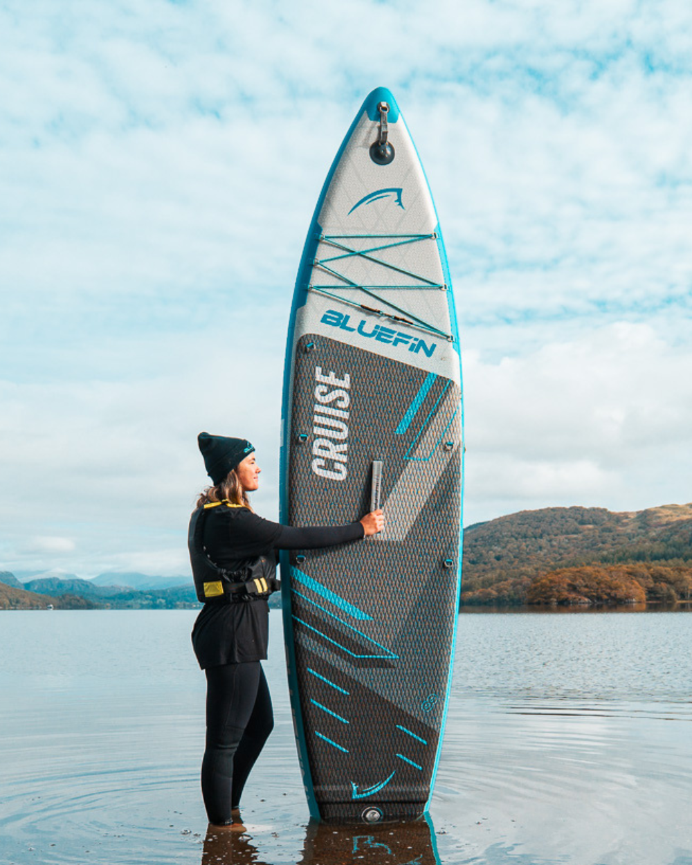 Woman in black outfit holding a Bluefin inflatable paddleboard in shallow water with mountains in background