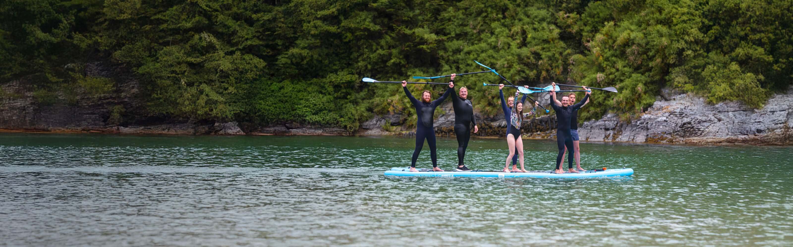 Group of six people on a paddleboard, enjoying a fun day on the water in wetsuits.