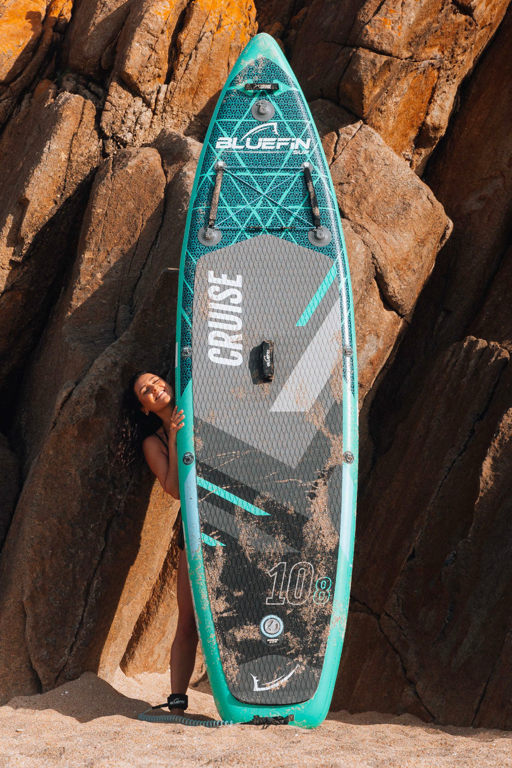Woman joyfully poses with a Bluefin SUP paddleboard against rocky beach backdrop.