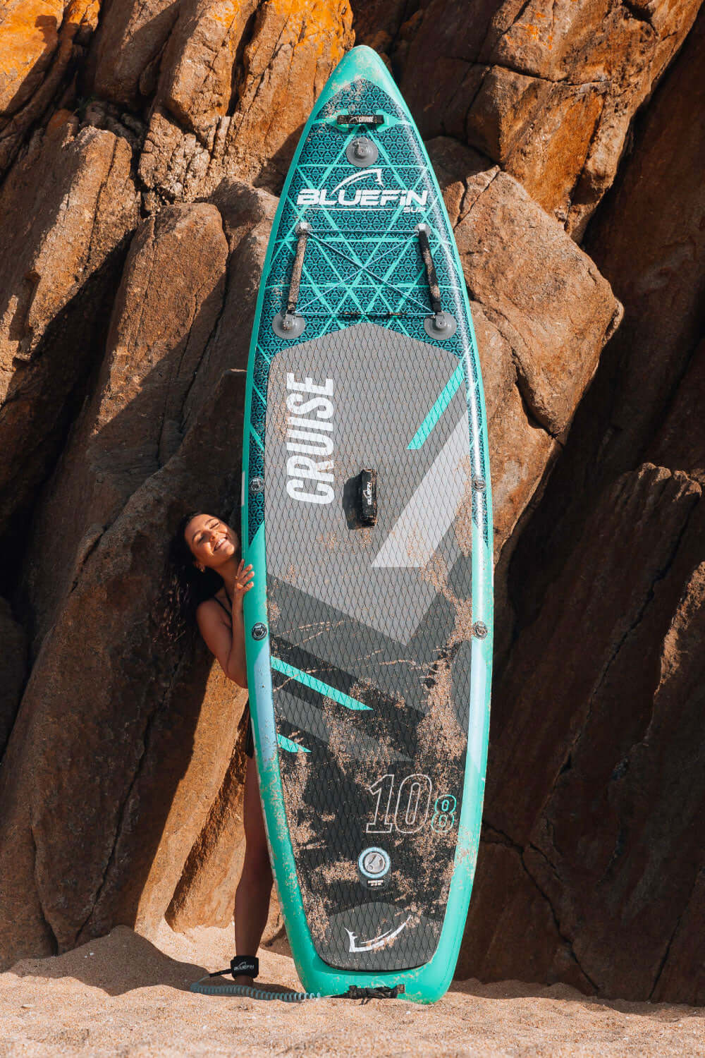 Woman joyfully poses with a Bluefin SUP paddleboard against rocky beach backdrop.
