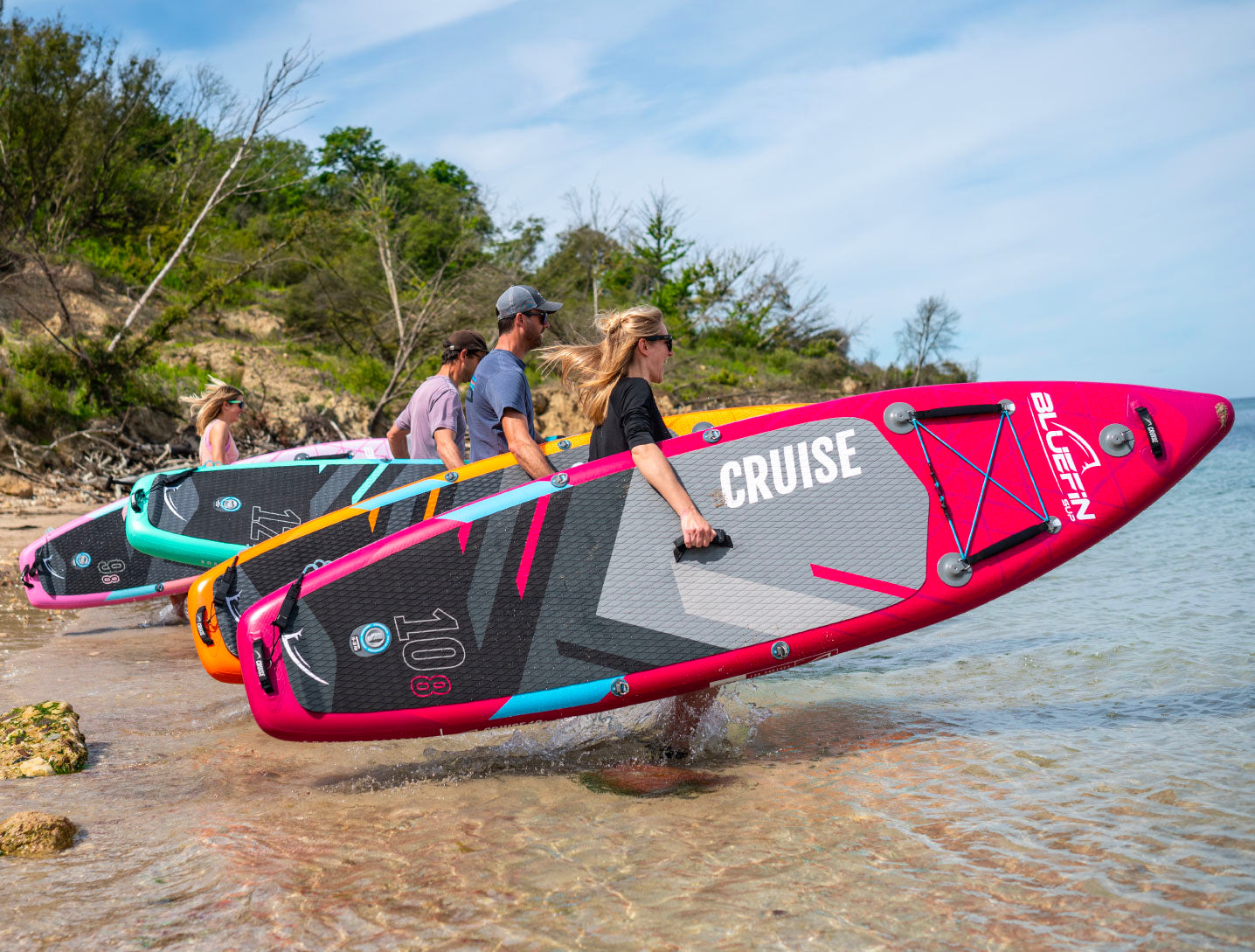 Group carrying colorful Bluefin SUP boards along the shore on a sunny day. Ideal for outdoor adventure.