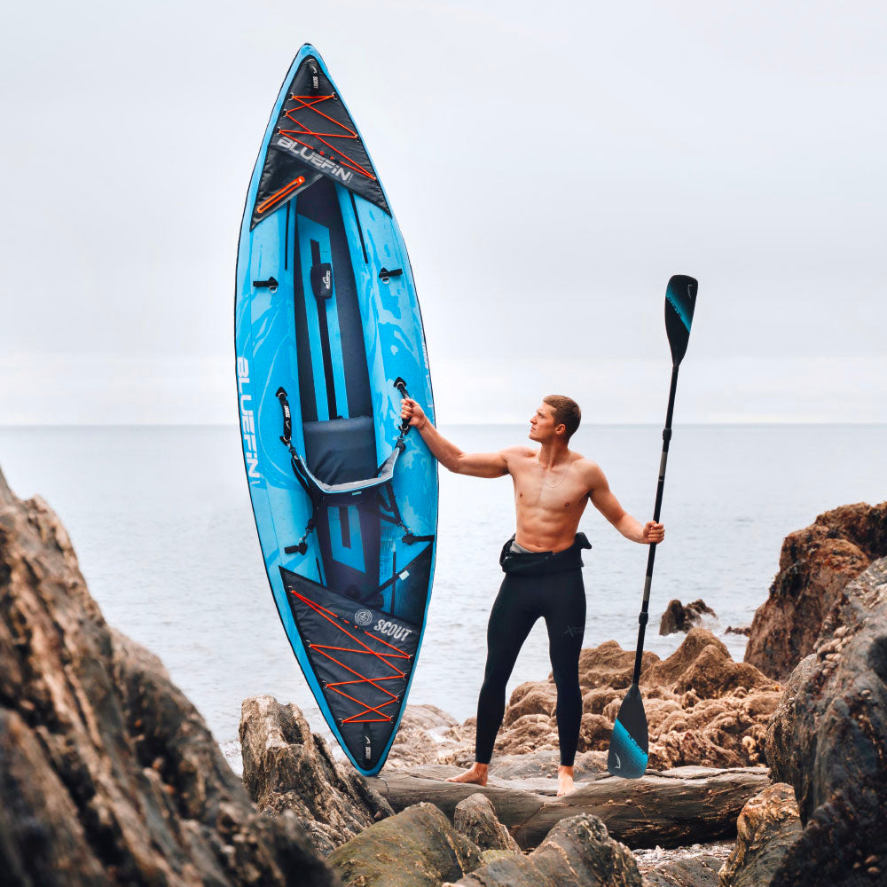Man holding a Bluefin stand-up paddleboard on rocky shoreline, ready for adventure.
