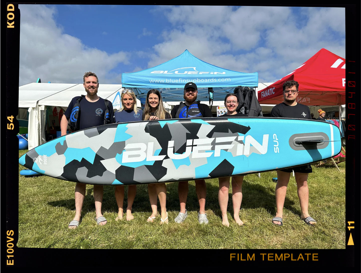 Five people smiling with a Bluefin SUP paddleboard, set against a sunny outdoor event backdrop.