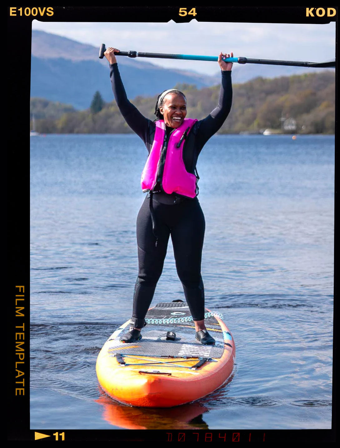 Woman celebrating on a paddleboard in a bright pink life jacket on a quiet lake.