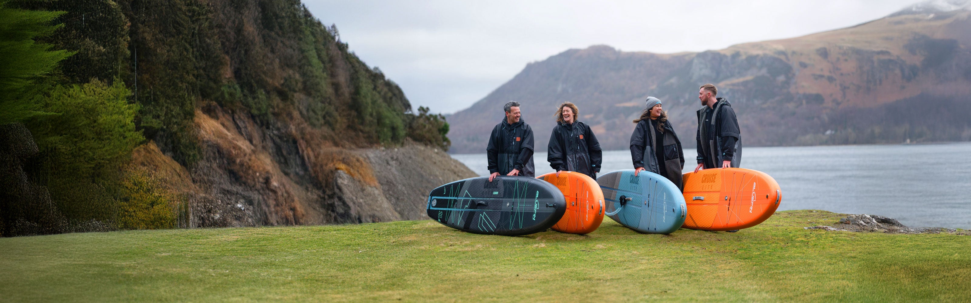 Group of four people standing on grass with colorful SUP boards and scenic lake backdrop. Ideal for paddling enthusiasts.
