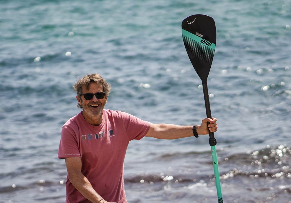Smiling man in sunglasses holds a paddle by the water, enjoying stand-up paddleboarding.