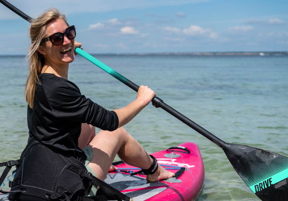 Woman smiling while paddling on a pink SUP board in clear water, wearing sunglasses and casual attire.