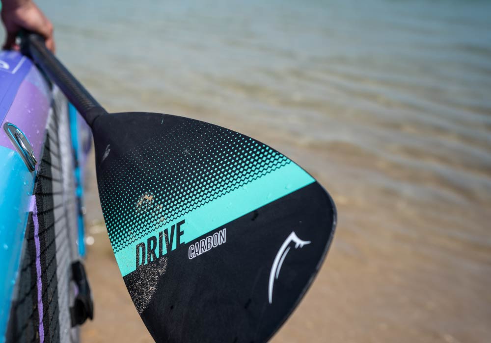 Carbon paddle close-up with "DRIVE" branding, against a sandy beach backdrop.
