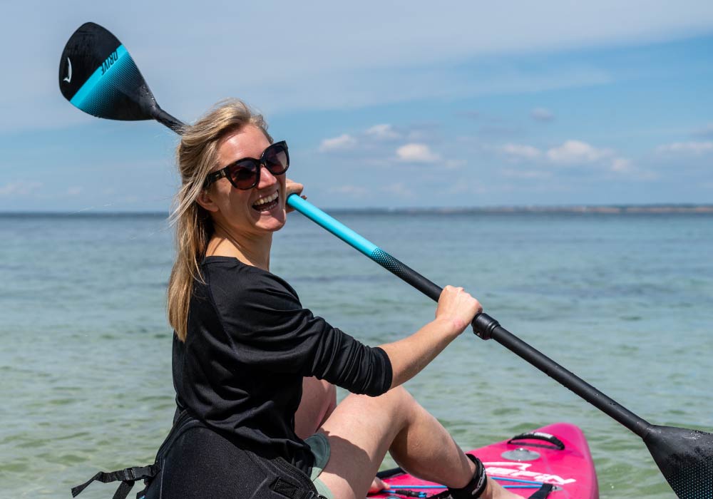 Smiling woman on a pink paddleboard in shallow water, holding a paddle against a sunny sky.