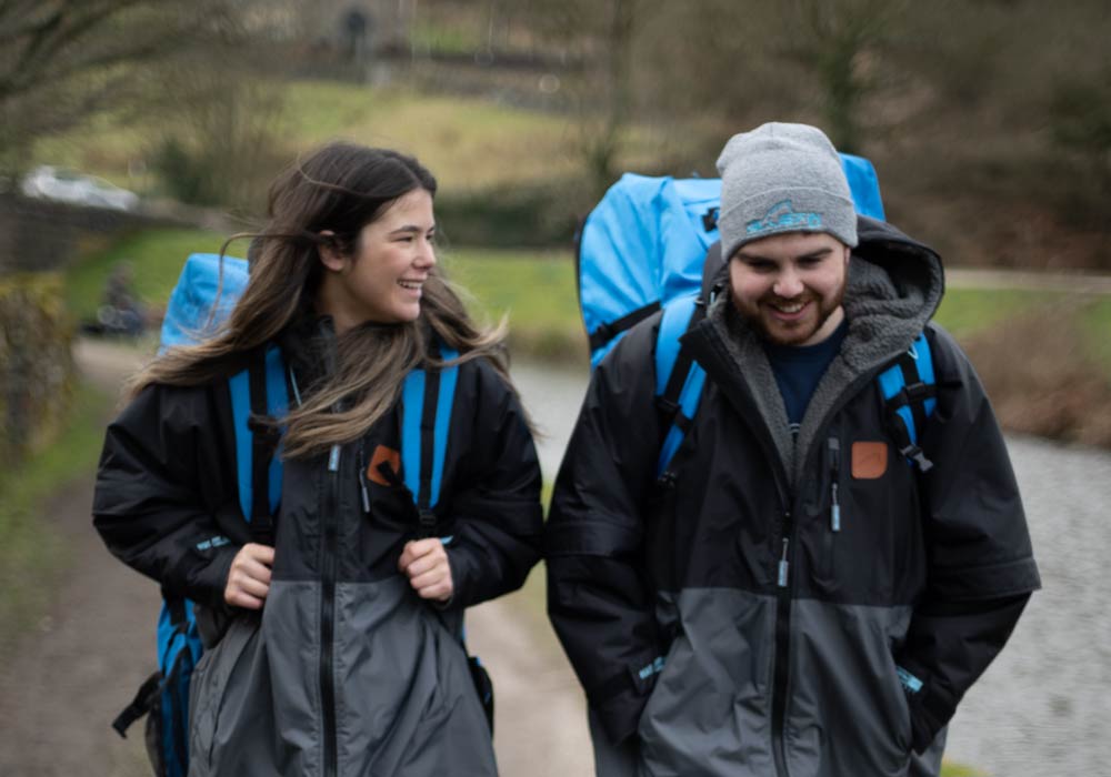 Two hikers in outdoor gear walk along a path by a waterway, smiling and enjoying their adventure.