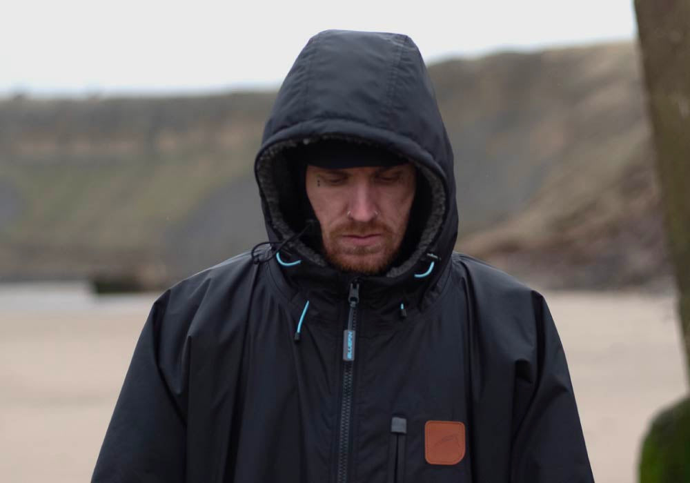 Man in a black jacket with hood, looking down on a beach, outdoors, cloudy atmosphere.