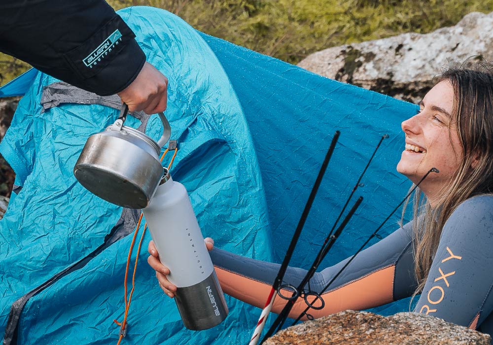 Happy person in wetsuit receiving a kettle while sitting by a tent outdoors. Enjoying nature and fishing.