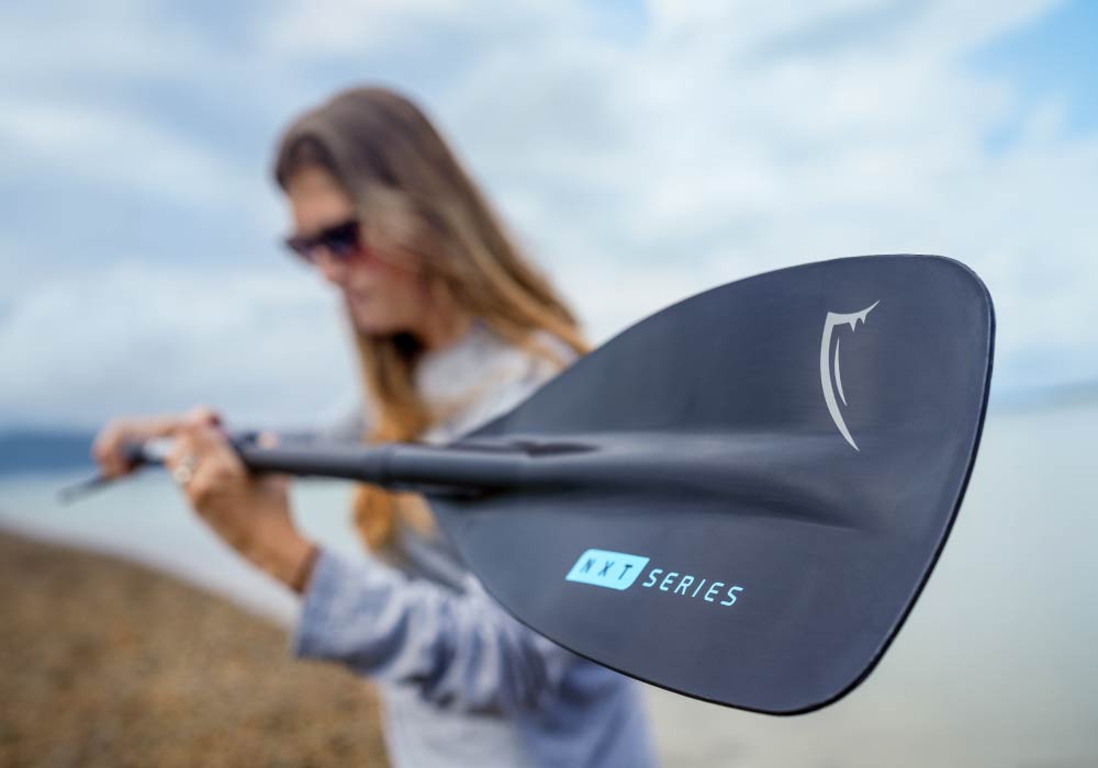 Woman holding a Bluefin SUP paddle on a beach, showcasing NXT Series design.