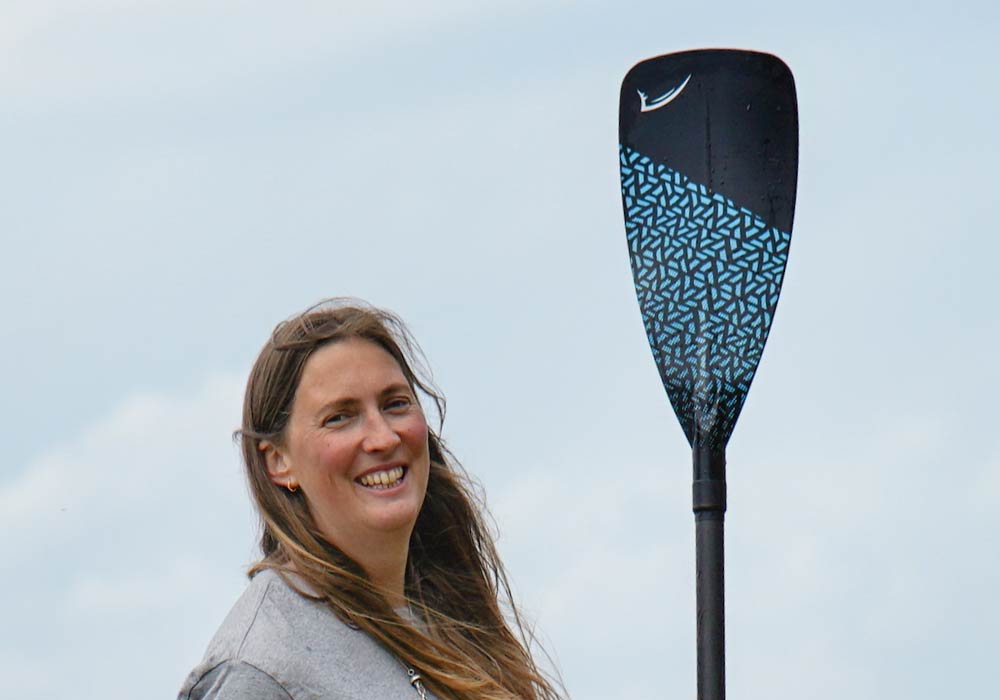 Smiling woman holding a paddle with a blue design against a cloudy sky backdrop.