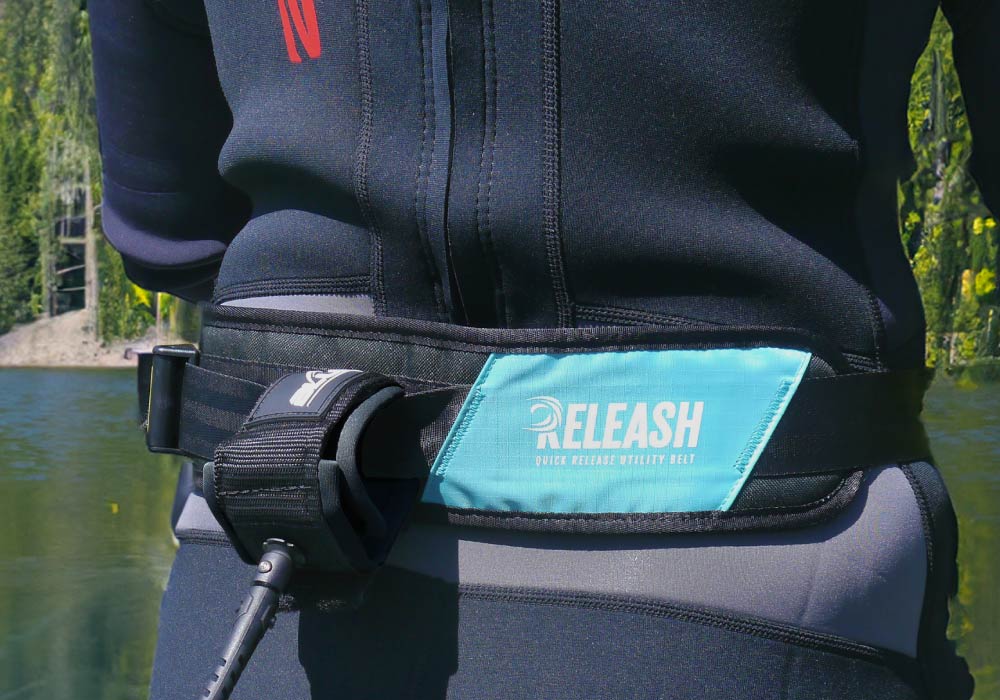 User wearing a wetsuit with a blue "RELEASH" quick release vitality belt near a body of water.