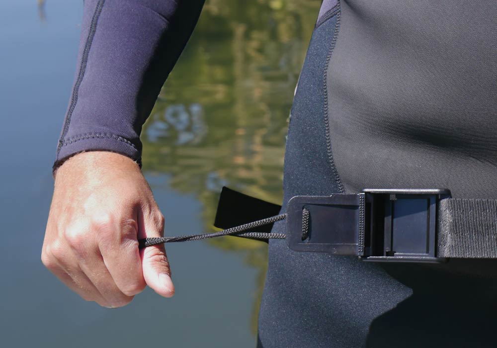 Person fastening a waist strap on a wetsuit by a calm body of water.