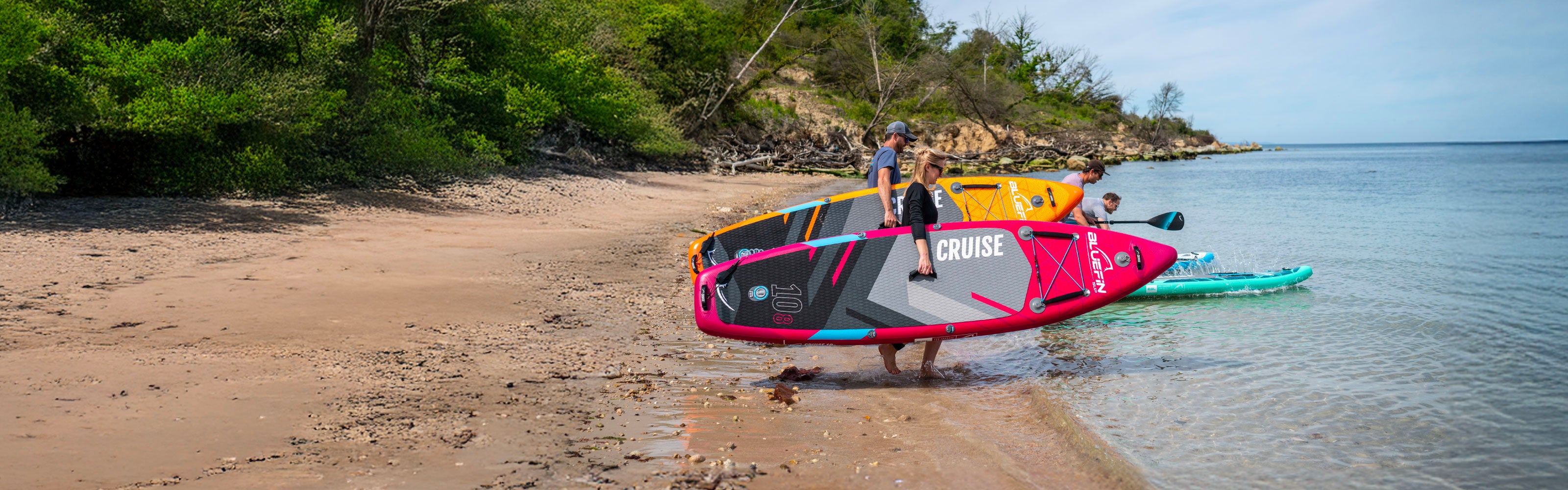Two paddle boarders carry colorful SUP boards along a sandy beach with lush greenery and calm water.