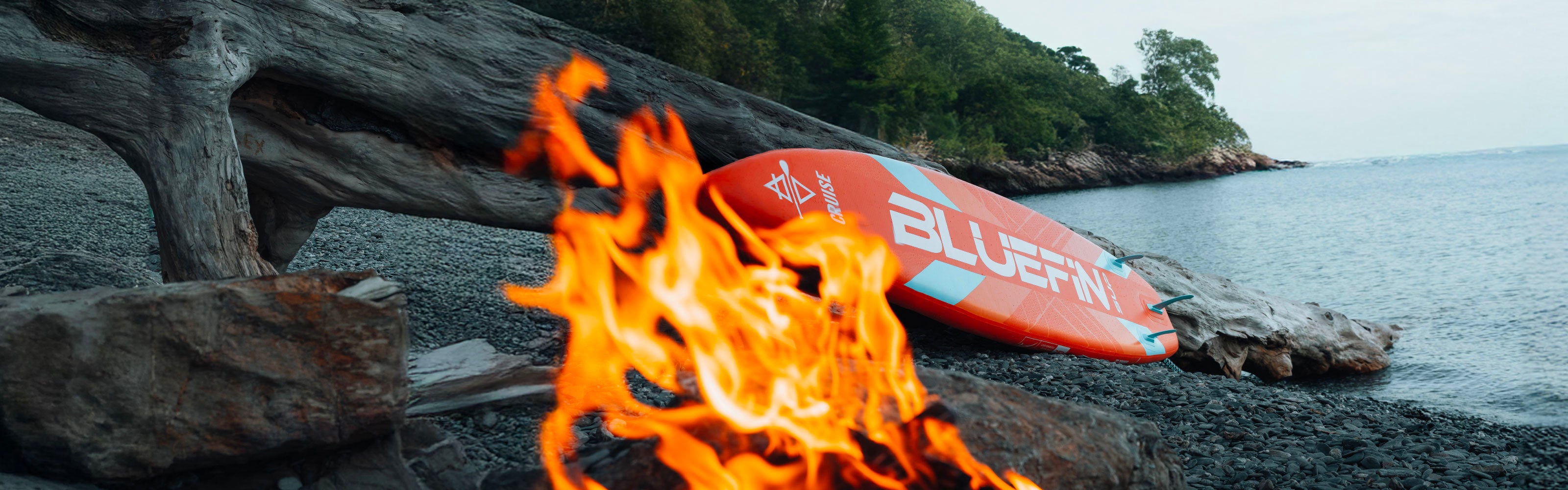 Orange Bluefin SUP board resting near a beach fire on a rocky shore with trees in the background.