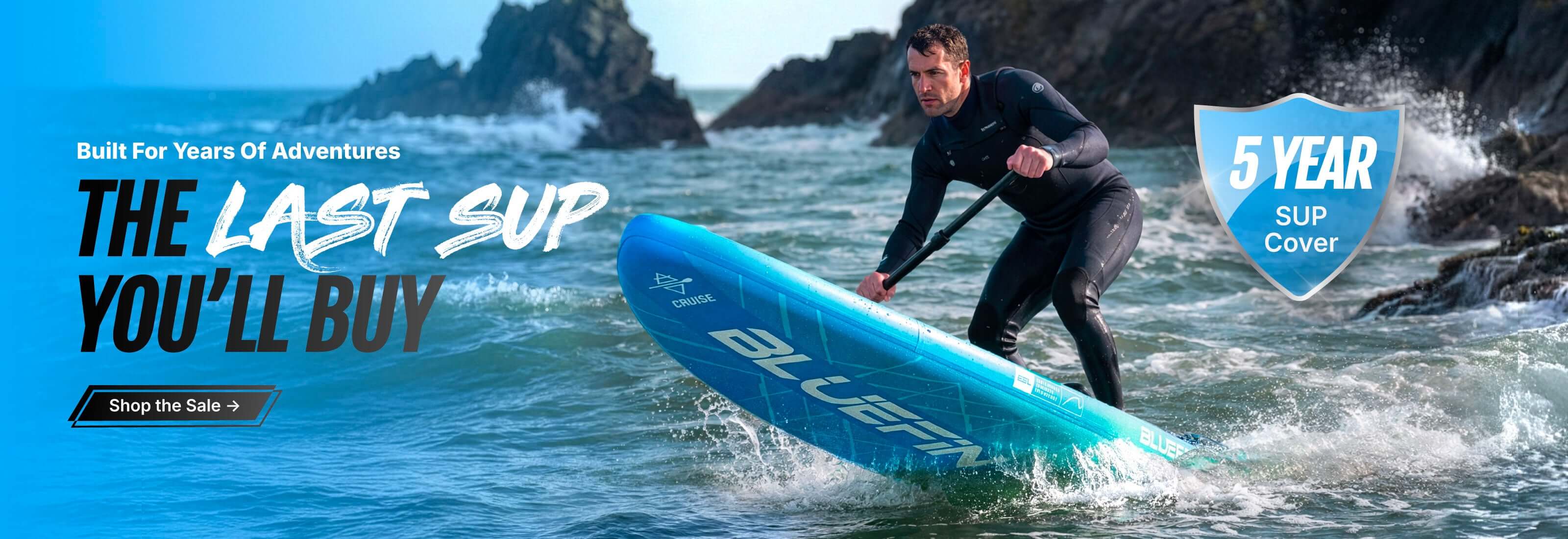 Man paddling a blue inflatable paddleboard in ocean water near rocky shore
