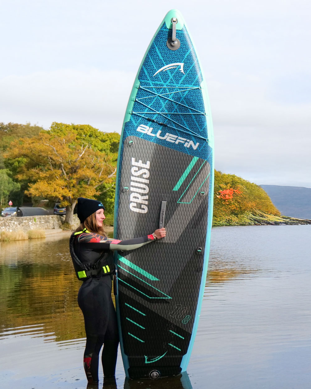 Woman in wetsuit holding a Bluefin inflatable paddleboard with textured surface and center fin in shallow water.