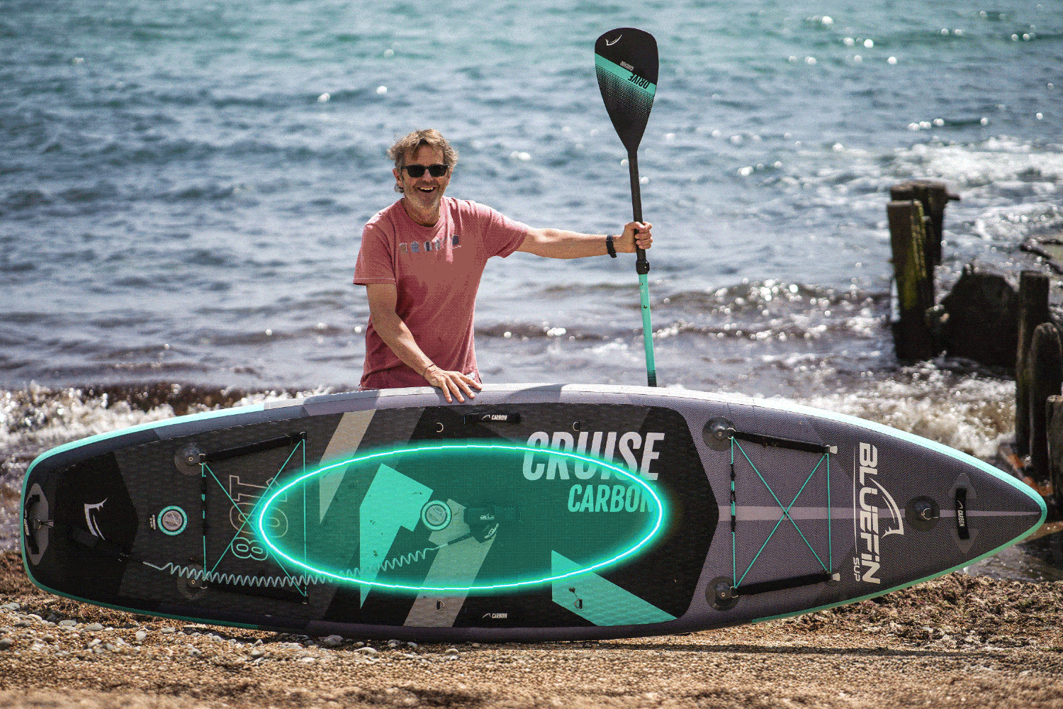 Man posing with a Bluefin SUP board and paddle on a beach by the water.