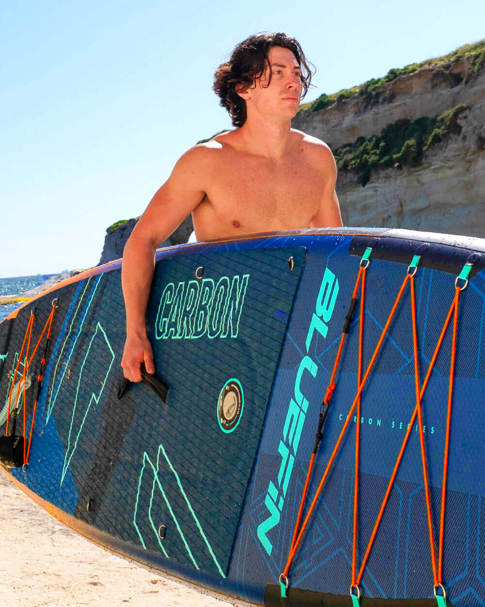 Man carrying a Bluefin Carbon Series paddleboard with textured surface and orange bungee cords on beach