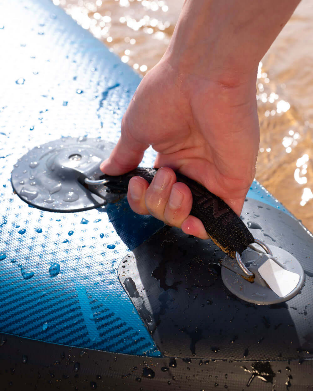 Hand gripping the handle of a blue inflatable paddleboard with water droplets on the surface