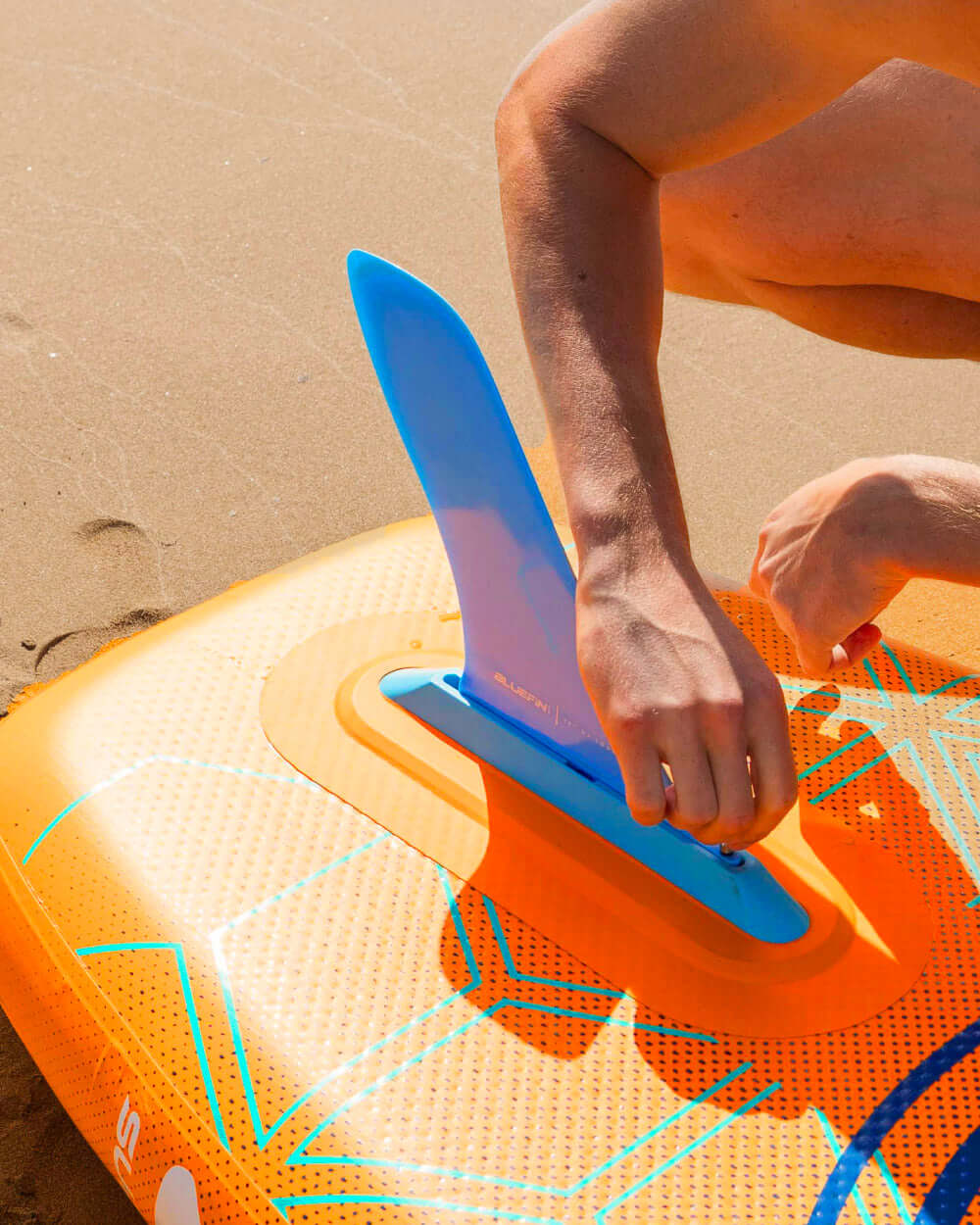 Person attaching a blue fin to an orange inflatable paddleboard on sandy beach