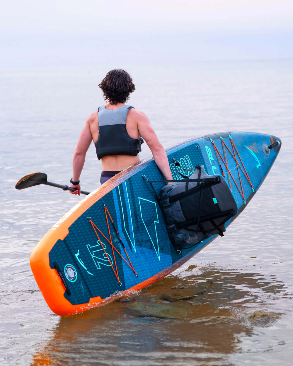 Person carrying a blue inflatable paddleboard with orange accents and a backpack, walking into shallow water