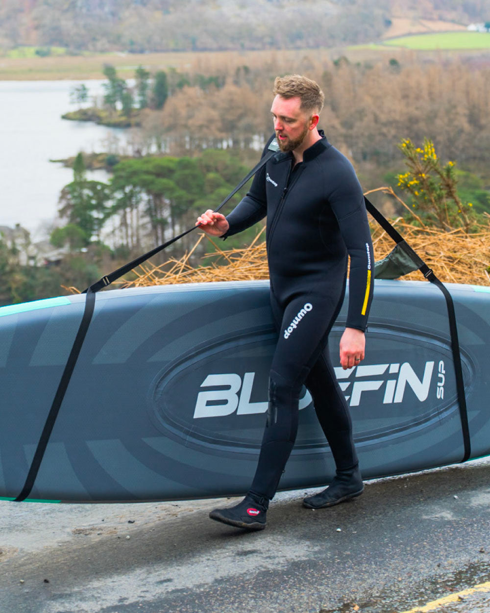 Man in a wetsuit walking with a Bluefin SUP board on a road near a lake using the carry strap designed for stand up paddle boards and kayaks