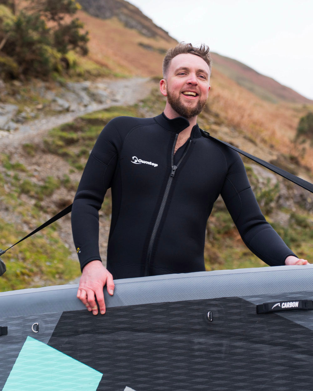 Man in a wetsuit holding an inflatable SUP board with mountains in the background using the sup carry strap