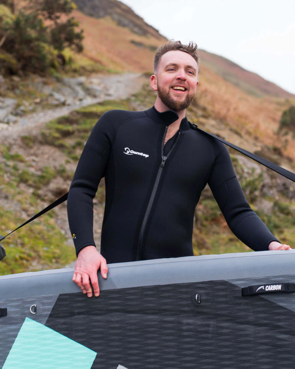 Man in a wetsuit holding an inflatable SUP board with mountains in the background using the sup carry strap