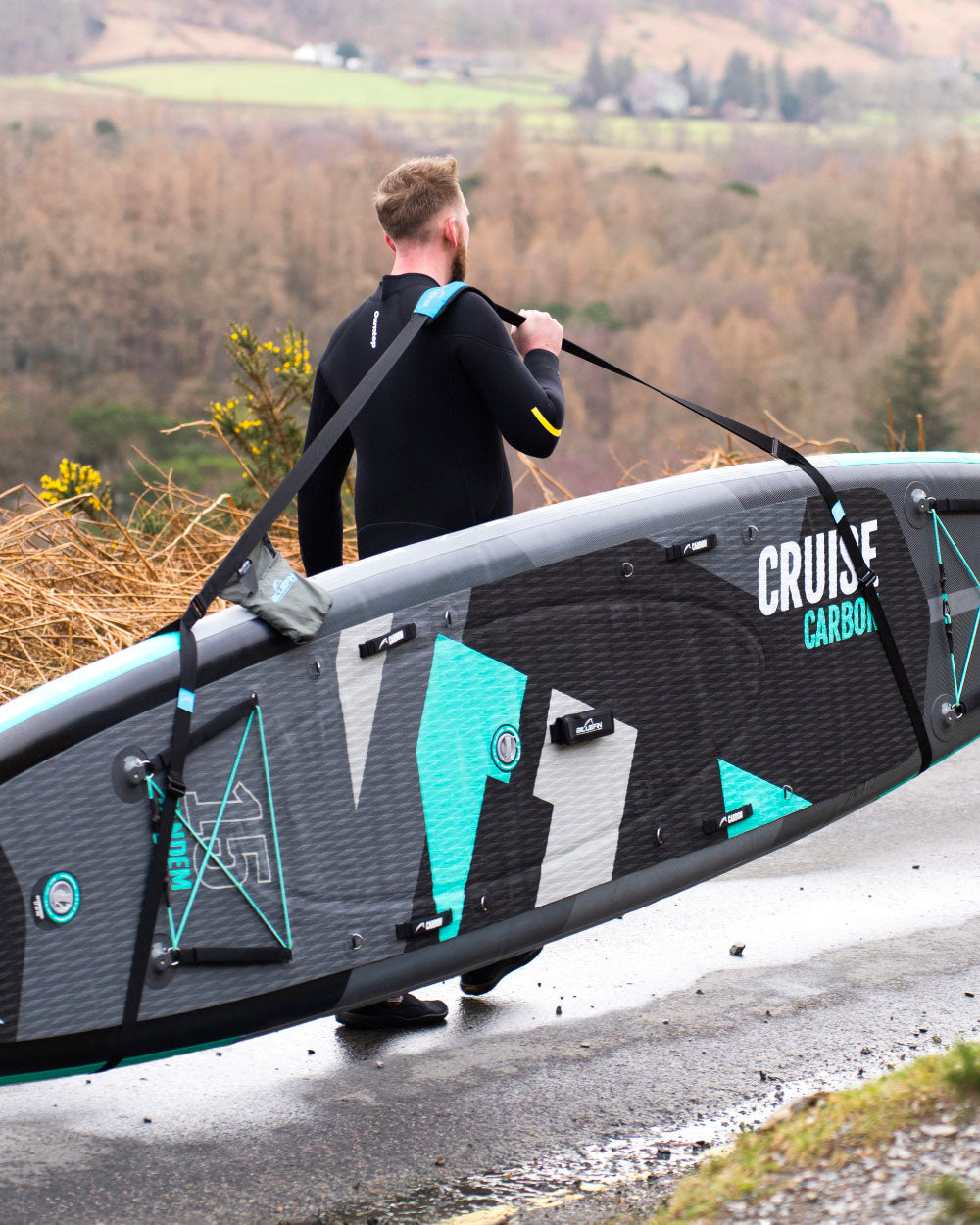 Person carrying a surfboard with 'Cruise' logo on a road with a scenic background using the sup carry strap