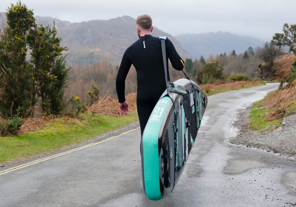 Person in wetsuit carrying a paddleboard along a scenic road with greenery and mountains in the background.