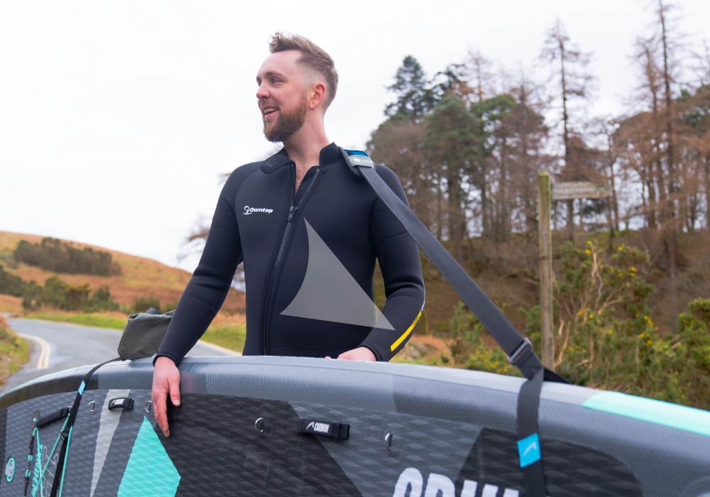 Man in a wetsuit standing beside a paddleboard on a scenic road.