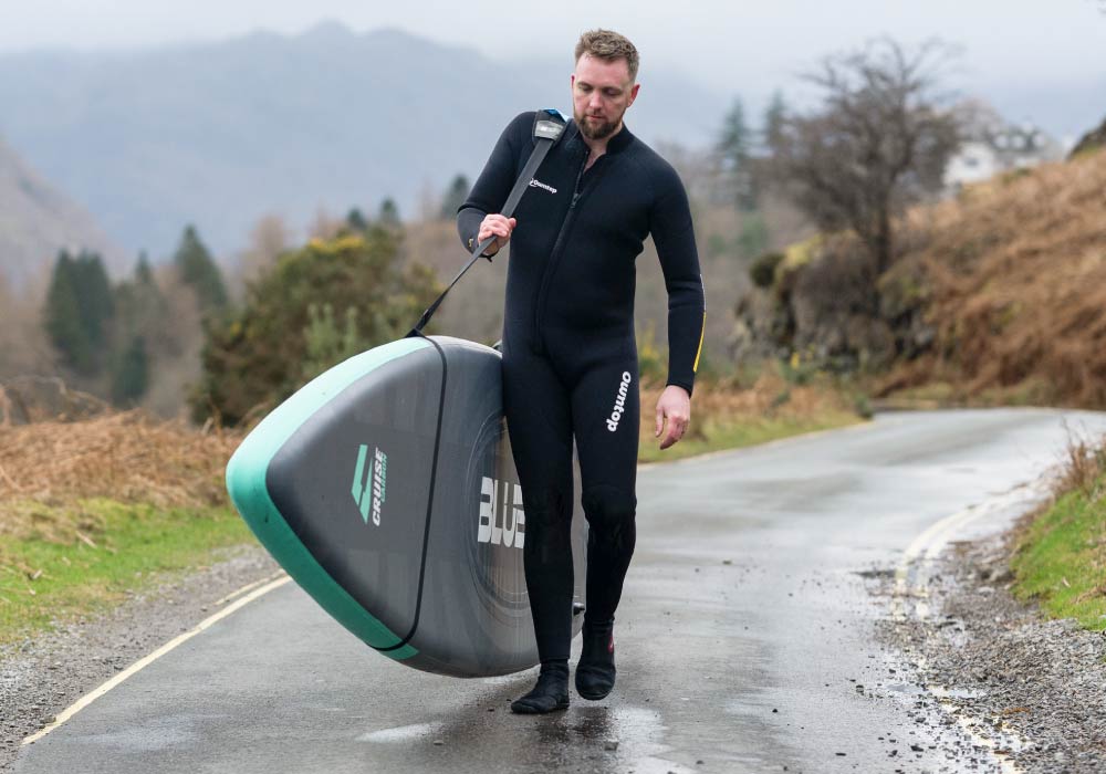 Person in a wetsuit carrying a paddleboard along a road in a scenic outdoor setting.