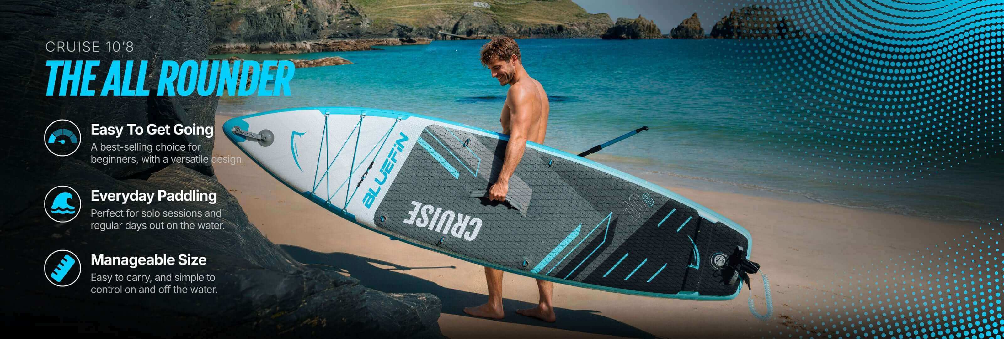 Man carrying a blue and gray inflatable paddleboard on a beach with rocks and water in the background