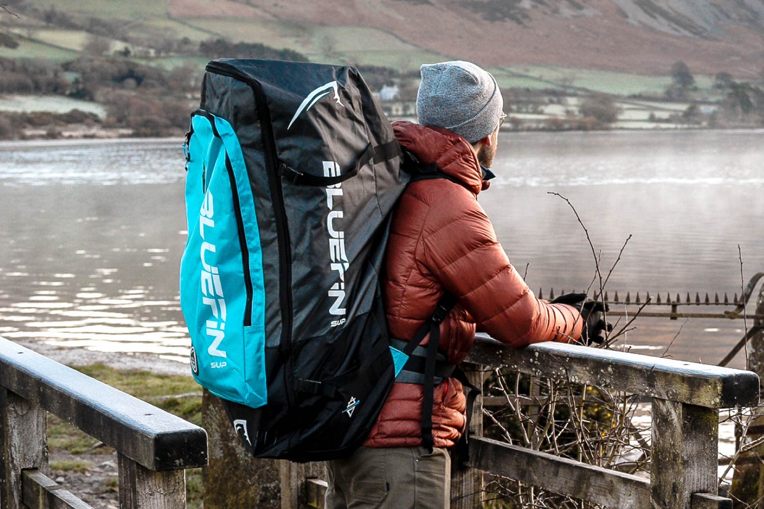 Person with Bluefin SUP backpack, gazing at a serene lakeside scene, enjoying outdoor adventure.