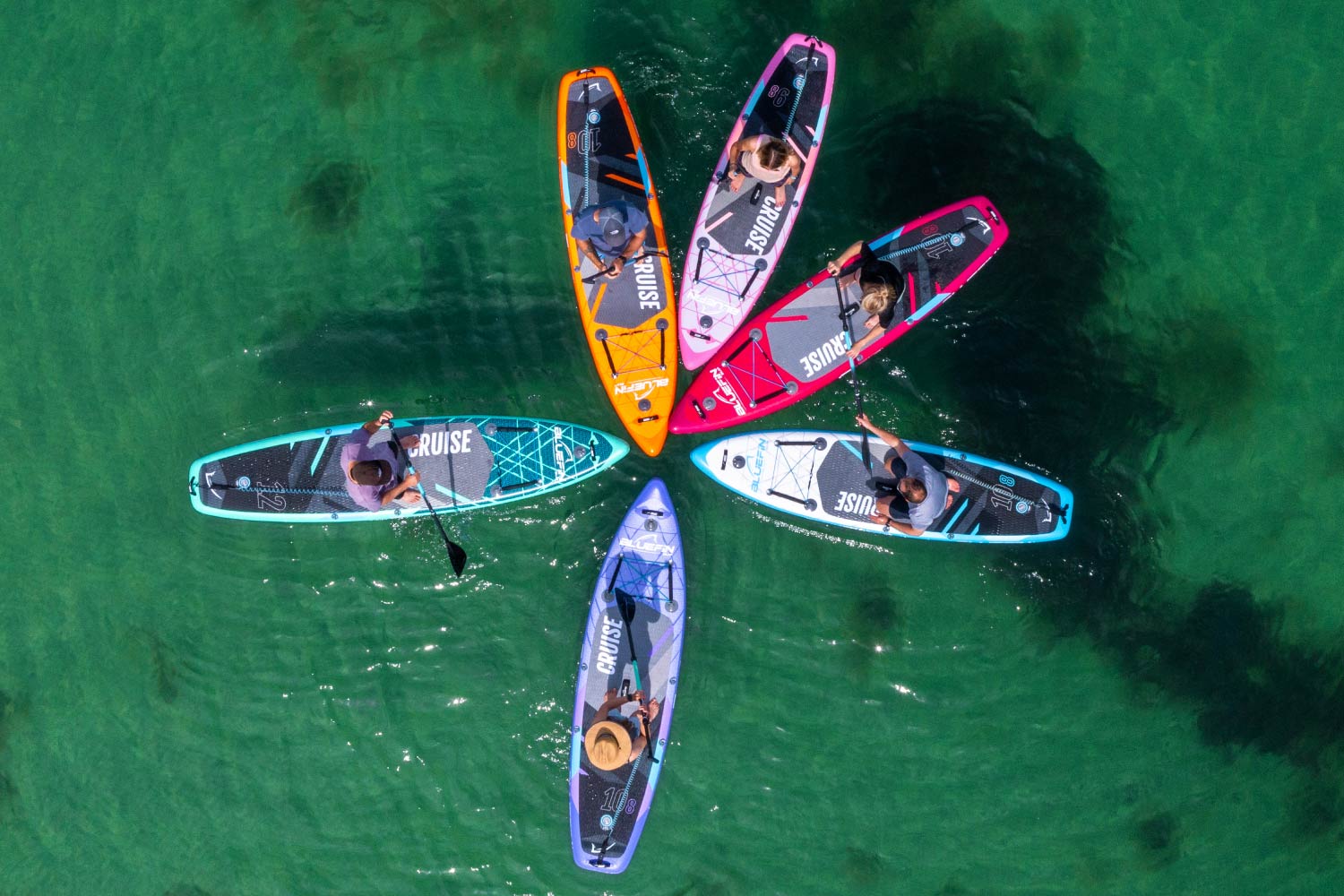 Group of paddleboarders in colorful boards arranged in a circular formation on clear water.