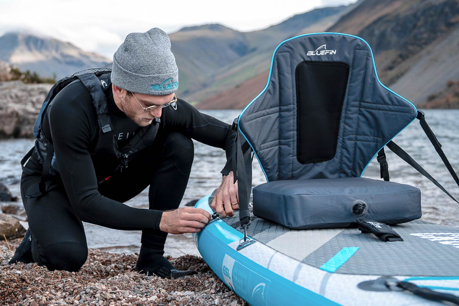 Man in wetsuit adjusting a Bluefin SUP paddleboard by water. Features a comfortable backrest.