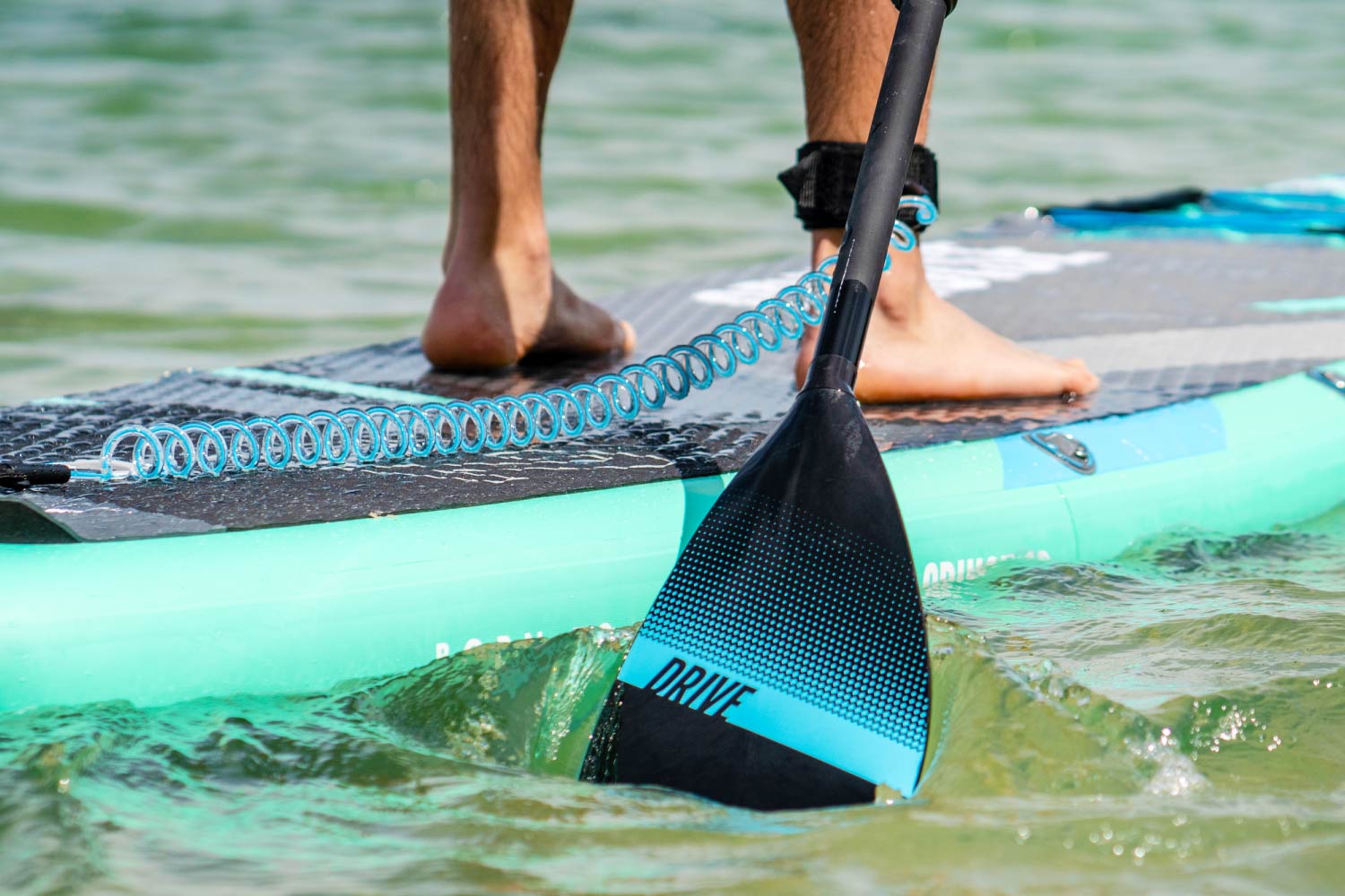 A person paddles a turquoise SUP with a paddle in shallow water, emphasizing an active water sport.