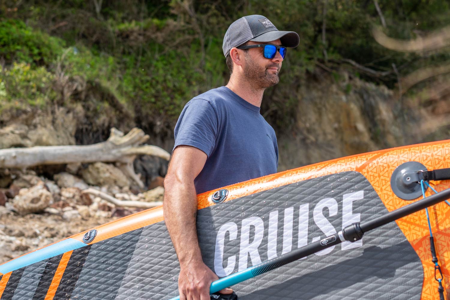 Man holding a paddleboard with "CRUISE" text, wearing sunglasses and a cap, on a beach background.