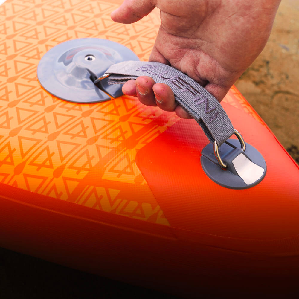 Hand holding the strap of a bright orange Bluefin SUP paddleboard with a patterned surface.