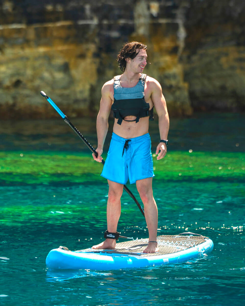 Man paddleboarding on a blue stand-up paddleboard in clear water, wearing a life vest and swim shorts.