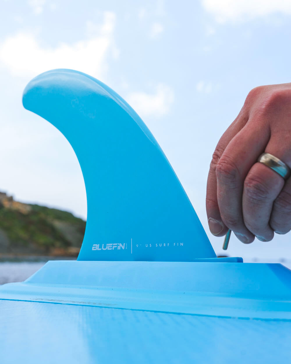 Bluefin SUP 9" US surf fin being installed on a paddleboard with a scenic background.
