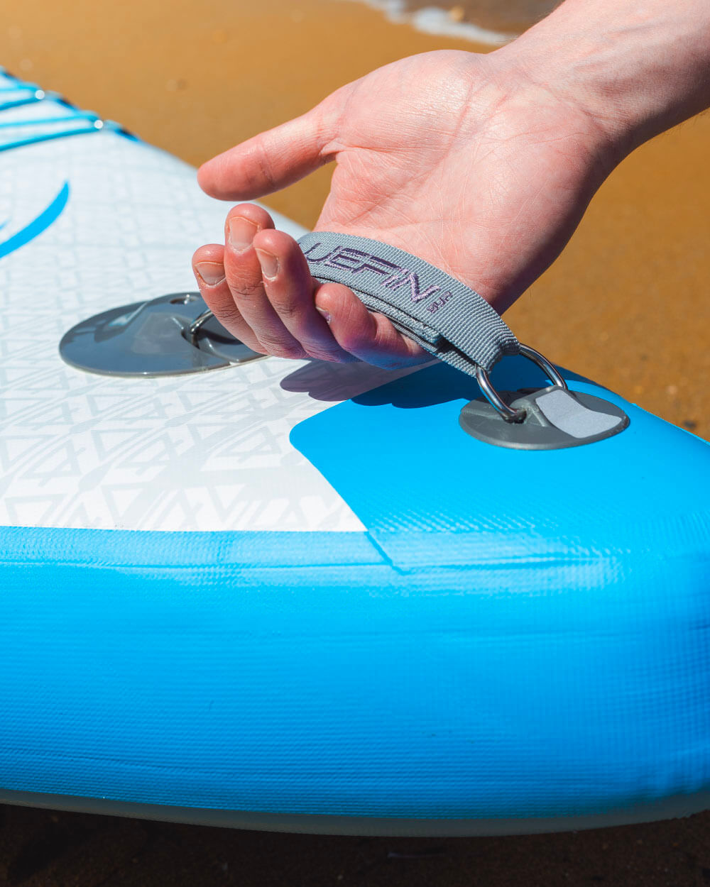 Hand holding a strap on a Bluefin paddleboard against a sandy beach background.