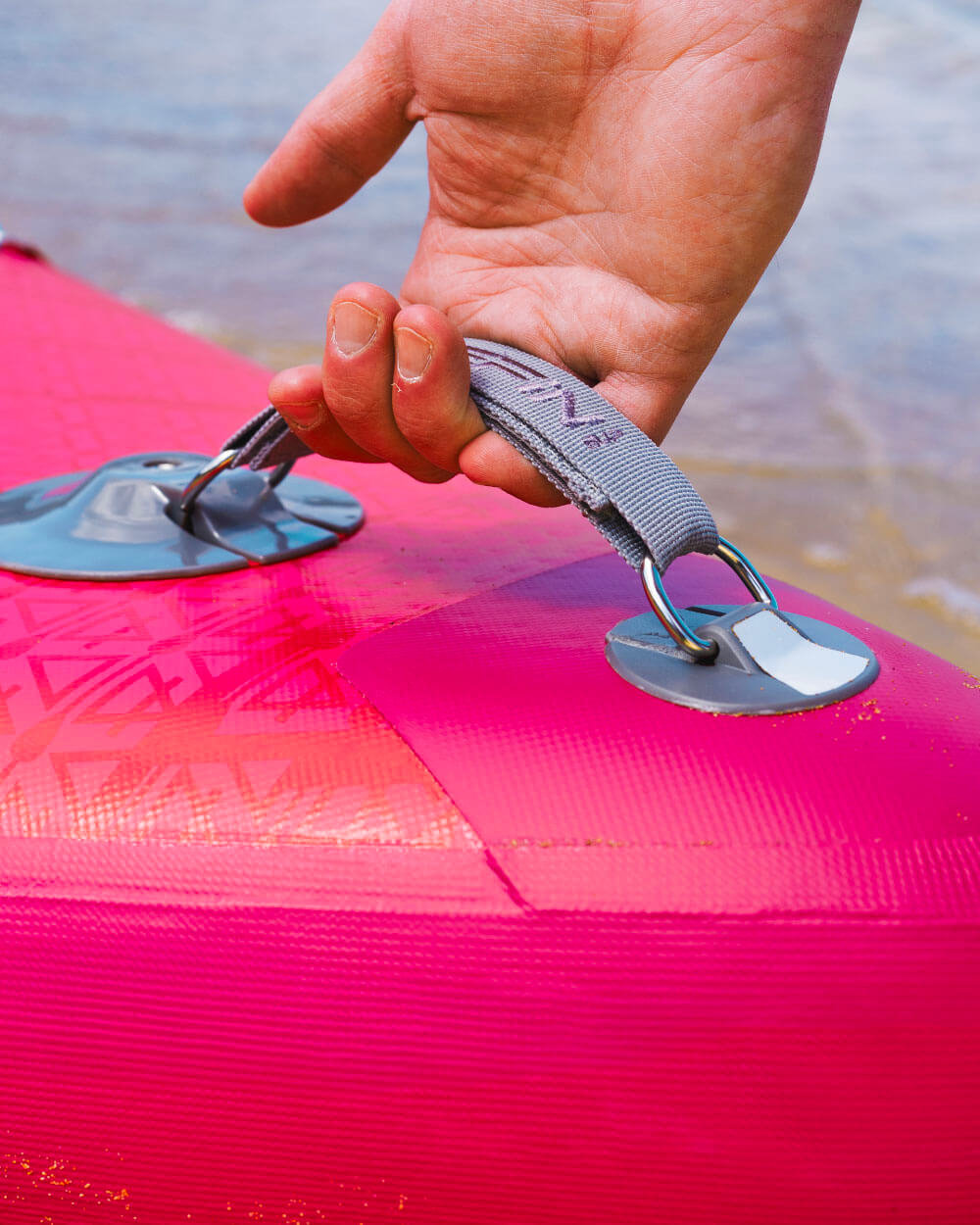 Hand holding a strap on a vibrant pink inflatable paddleboard by the water's edge.