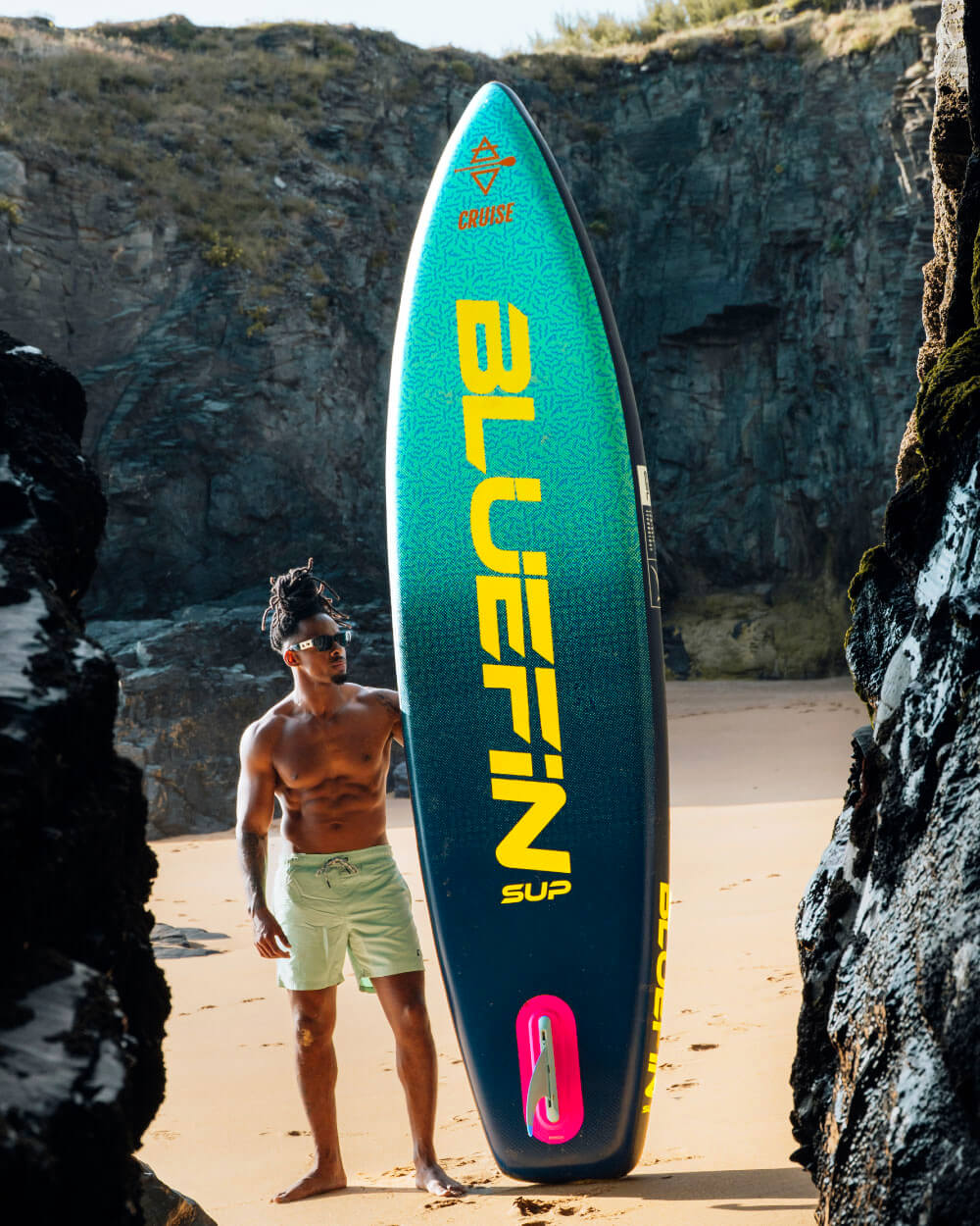 Man standing on a beach with a Bluefin SUP paddleboard against rocky cliffs, wearing sunglasses and shorts.